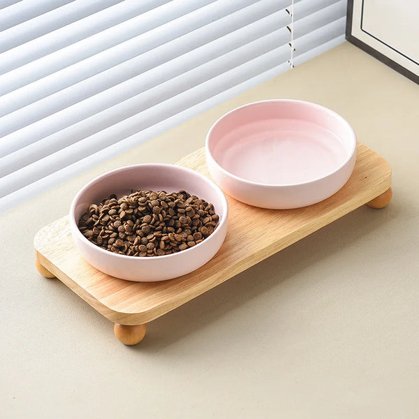 A pink ceramic cat and dog bowl is placed on a wooden stand next to a window blind.
