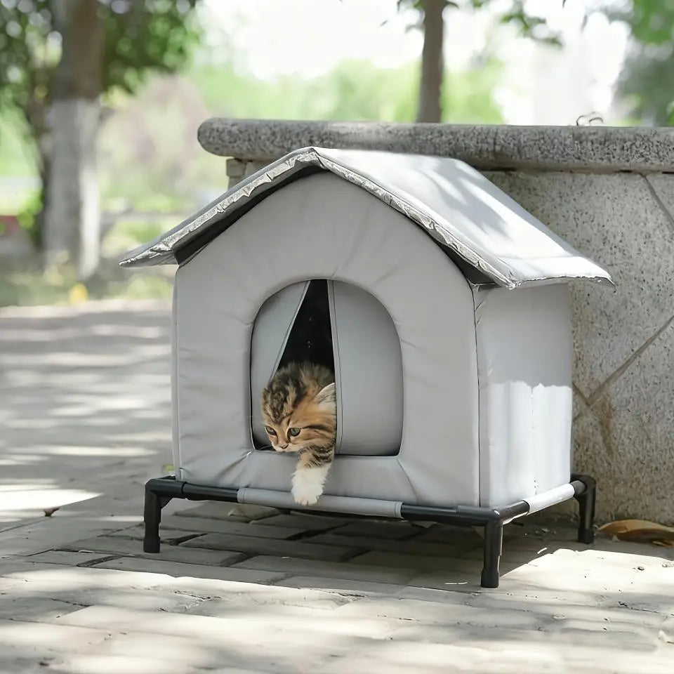 A gray outdoor cat shelter with a pitched roof is placed on a stone surface, with a tabby cat looking out.