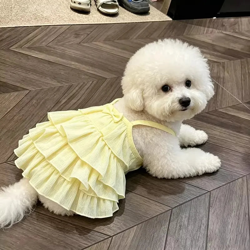 A fluffy white dog wearing a yellow ruffled summer strap dress lies on a wooden floor.
