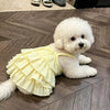 A fluffy white dog wearing a yellow ruffled summer strap dress lies on a wooden floor.