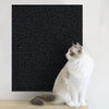 A white and gray cat is sitting on a wooden surface in front of a black textured mat.