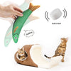 A hand is holding a green plush fish toy with an orange tail, next to a tabby cat.