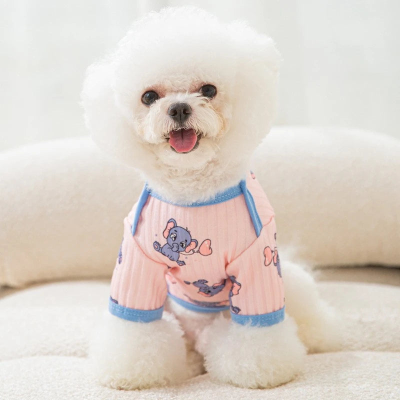 A white dog wearing a pink cartoon print vest, lying down on a light surface.