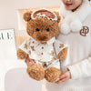 A child holds a brown teddy bear plush with pajamas and an eye mask against a white background.