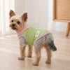 A small, fluffy dog wearing a light green and pink cartoon dress standing indoors.