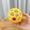 A hand holds a yellow natural rubber interactive dog chew ball with a pink bell.