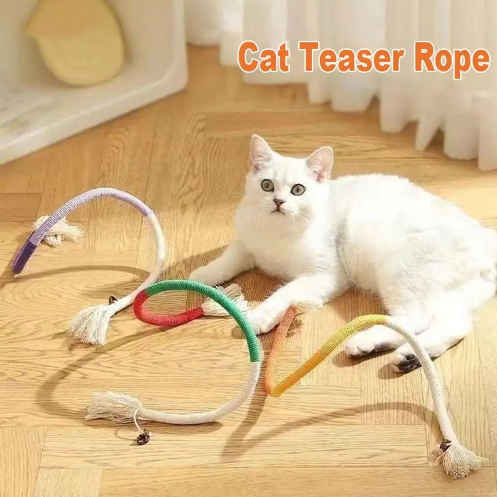 A white cat is playing with several colored cotton rope cat teaser toys on a wooden floor.