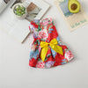 A vibrant red and pink floral print dog dress with a yellow bow, displayed on a white surface next to some greenery.