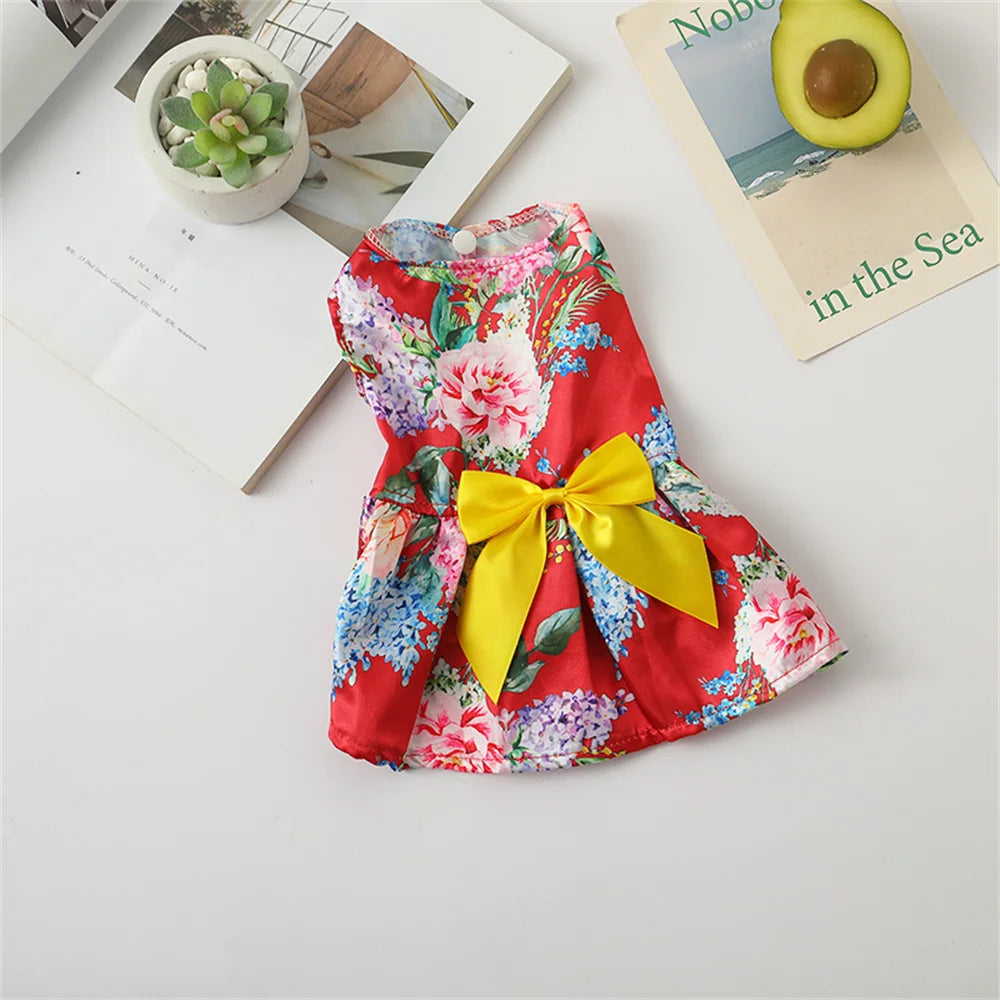 A vibrant red and pink floral print dog dress with a yellow bow, displayed on a white surface next to some greenery.