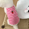 A white fluffy dog wearing a pink and white striped vest with a red strawberry print, sitting on a woven mat.