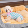 A light-colored poodle wearing a pink and green striped cotton recovery suit resting in a yellow pet bed.