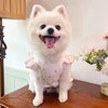 Un chien blanc et duveteux portant une robe d'été à motifs roses, assis sur une surface en bois.