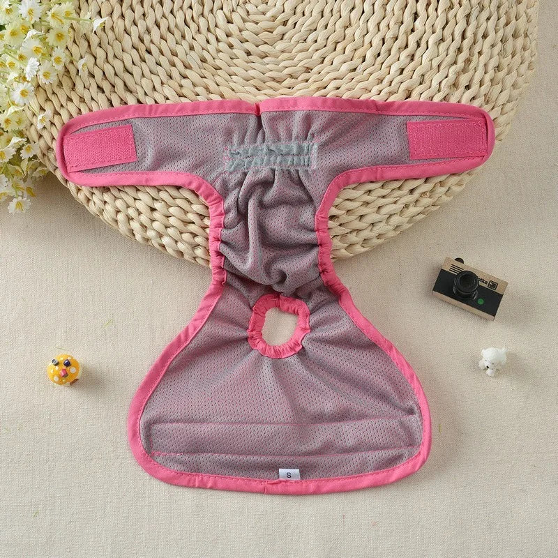 A pink and gray reusable dog diaper lying on a beige woven mat with a small camera nearby.