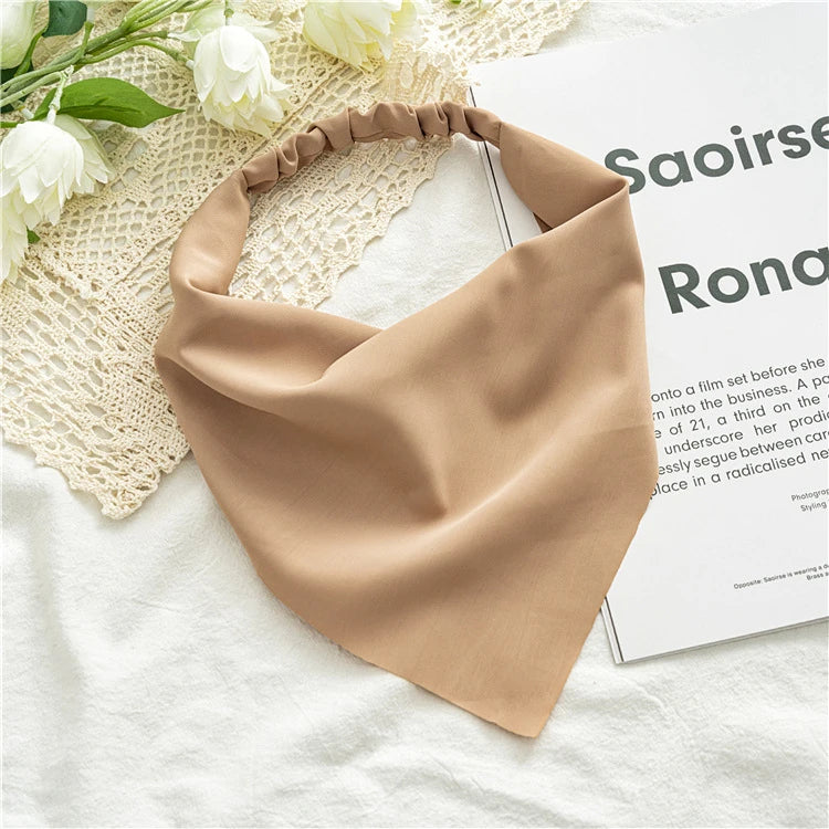 A beige fabric triangle head scarf is displayed on a white surface next to some flowers and a magazine.