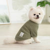 A fluffy white dog wearing a olive green knitted sweater sitting on a light surface.