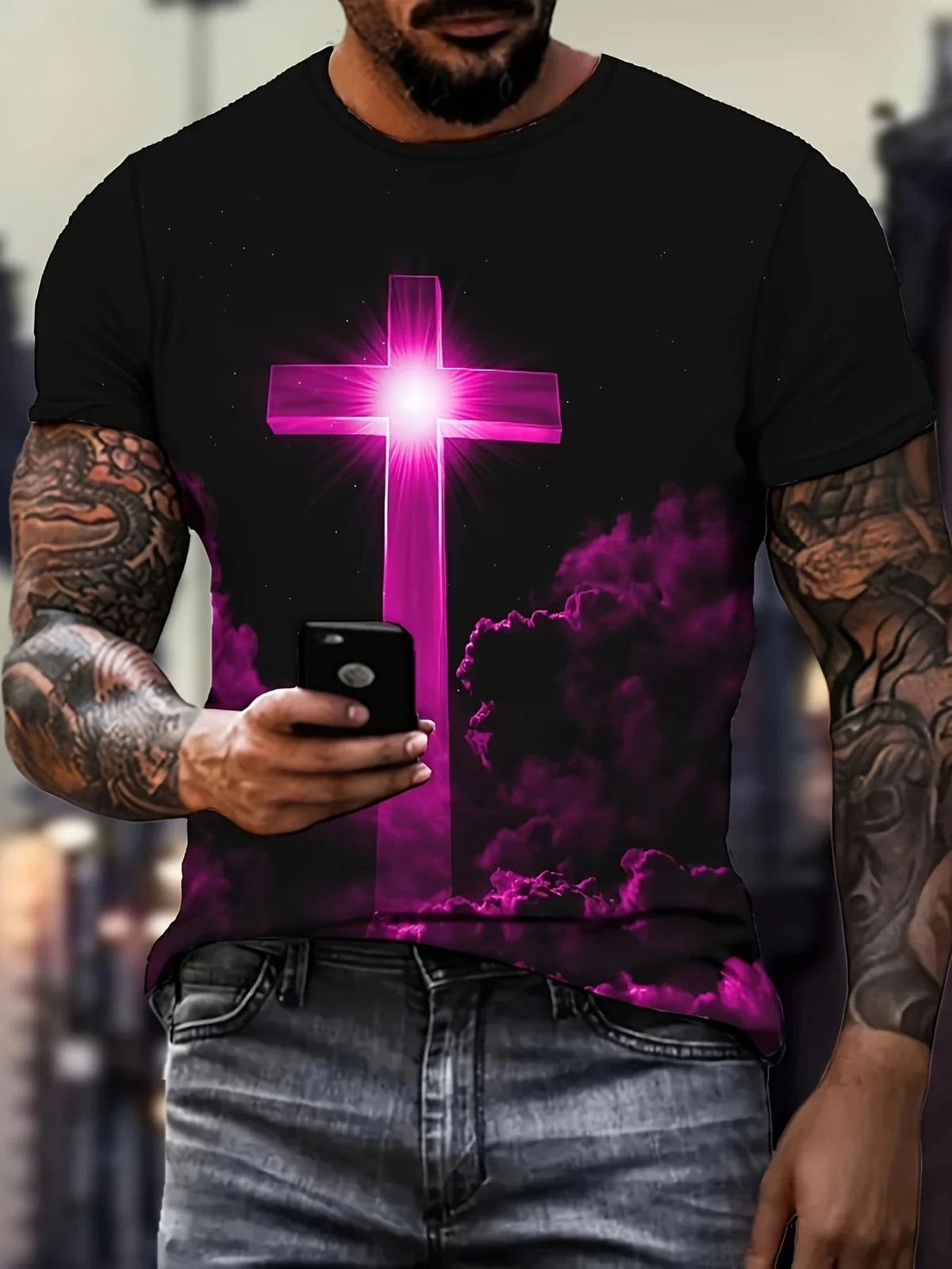 A man wearing a black t-shirt with a glowing pink cross design and dark clouds, holding a smartphone.