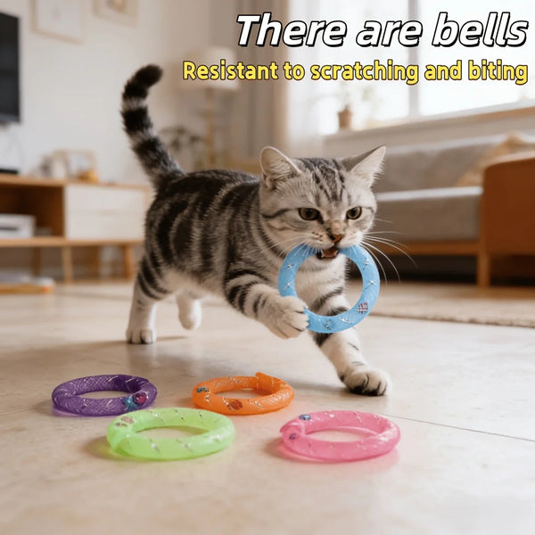 A tabby cat is playing with various colored donut-shaped chew toys, including a blue one with a bell.