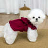 A fluffy white dog wearing a deep red vintage corduroy pet dress with a bow and ruffle skirt.