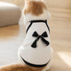 A fluffy orange and white cat wearing a white pet vest with a black bow knot.