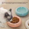 A cat is eating dry food from a pink ceramic pet bowl on a beige surface.