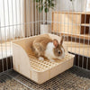 Un conejo pequeño marrón y blanco está sentado dentro de una caja de arena rejada beige desmontable en una jaula de alambre.