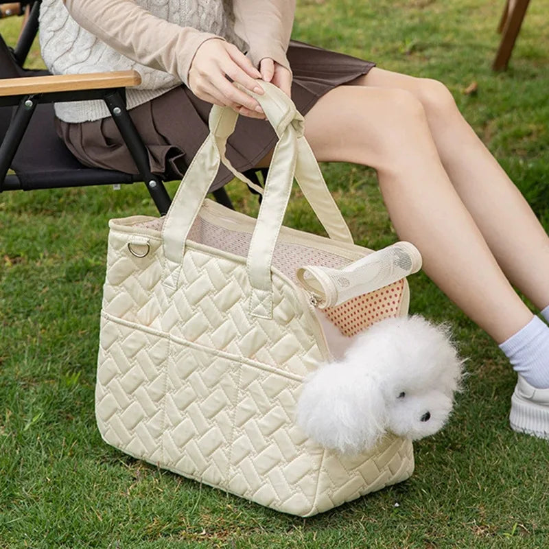 A cream quilted portable pet carrier bag with light beige handles rests on green grass next to a person.