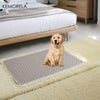 A golden retriever dog sits on a gray reusable pet training pad placed on a cream rug next to a wooden bed.
