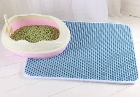 A pink and white double-layer cat litter mat with a green litter scoop resting on a blue textured mat.