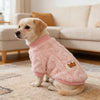 A light golden Labrador wearing a pink fleece pet sweater sitting on a beige rug indoors.
