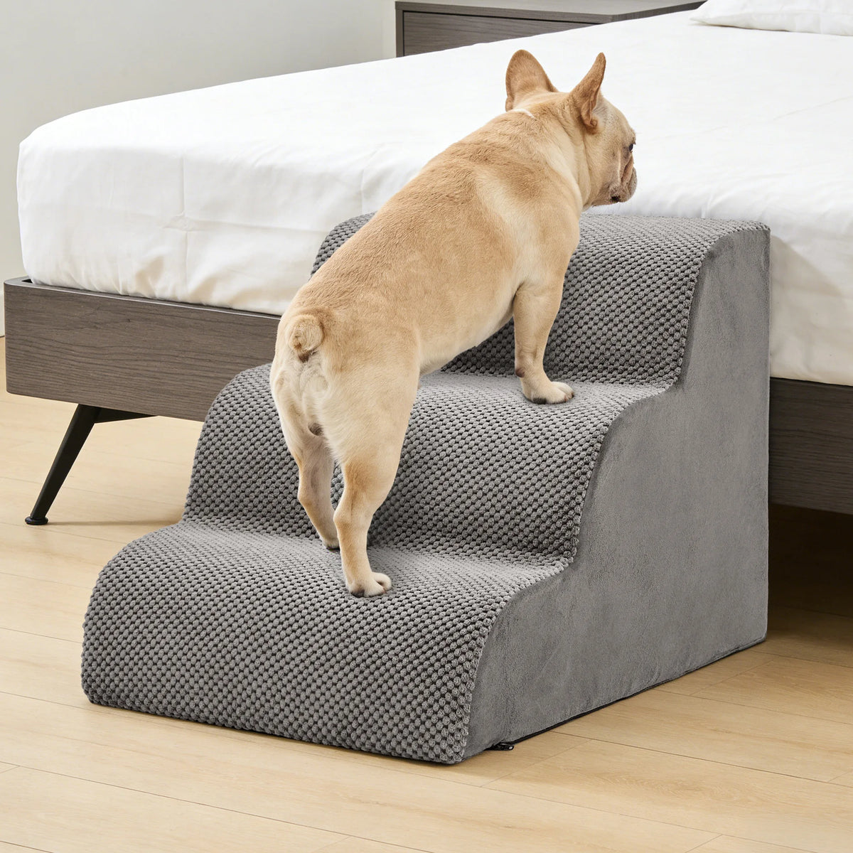 A light tan dog is stepping onto a gray foam pet stairs ramp placed next to a white bed.