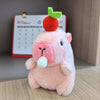 A pink capybara plush doll with green ears is sitting on a wooden surface.