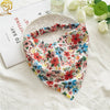 A floral patterned triangle head scarf is displayed on a light-colored surface next to a white ceramic dish.