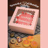 A pink box with a cutout design featuring red frosting text and decorative white icing, placed next to several round, glazed 