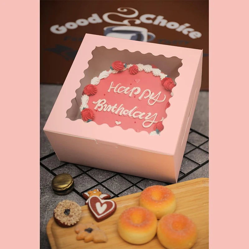 A pink box with a cutout design featuring red frosting text and decorative white icing, placed next to several round, glazed 