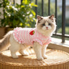Un gato blanco esponjoso vistiendo un vestido a rayas rosa y blanco con bordado de fresa, sentado sobre una superficie tejida