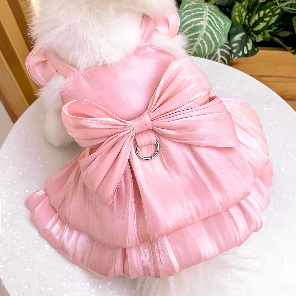 A pink satin pet dress with a bow and D-ring for a small dog is displayed on a white surface.