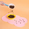 A hand is pouring dark pet food into a yellow bowl placed on a pink non-slip pet mat.