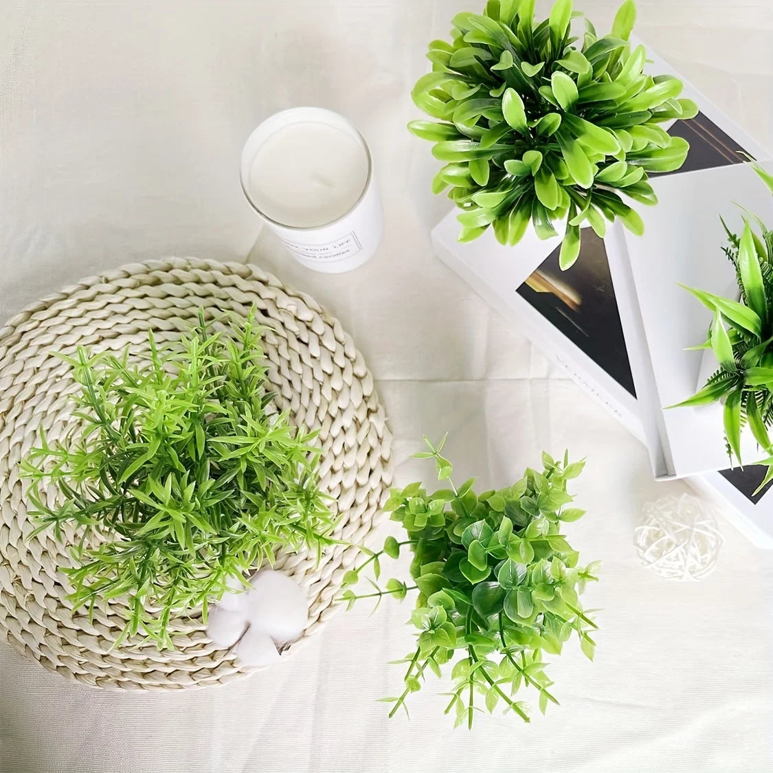 A collection of artificial bonsai potted plants displayed on a white surface with a candle and a woven mat.