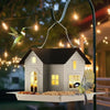 A solar outdoor bird feeder lantern with a house design is suspended by a metal wire on a dark pole.