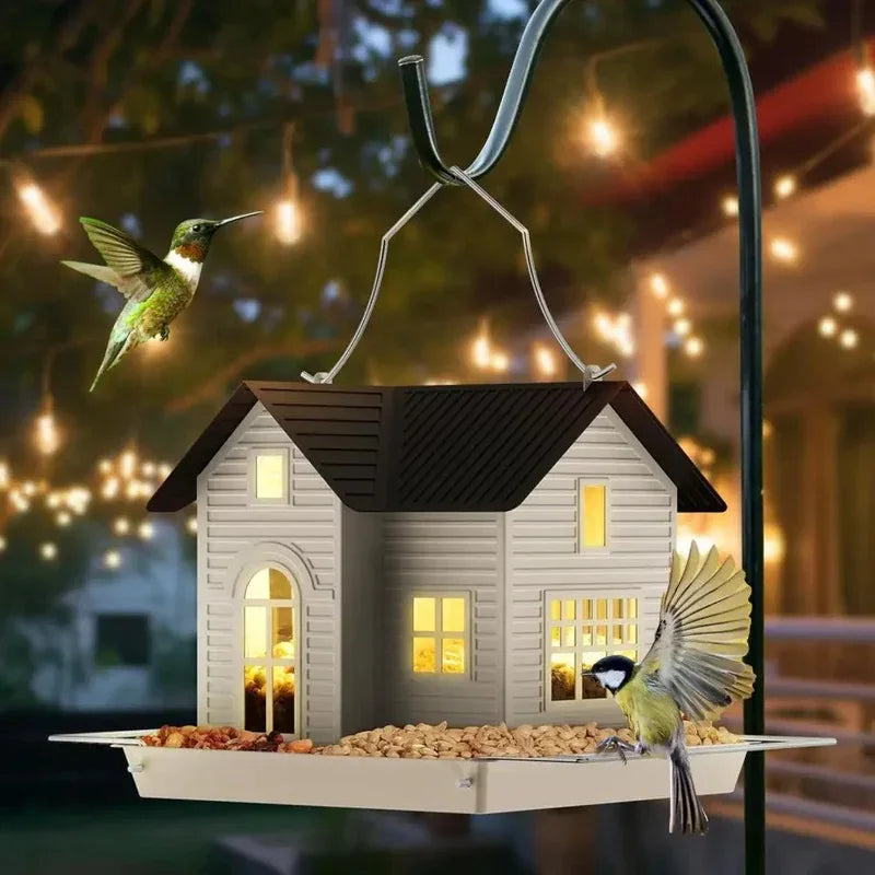 A solar outdoor bird feeder lantern with a house design is suspended by a metal wire on a dark pole.