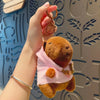 A hand holds a brown capybara plush doll wearing a pink hoodie and gold wings against a light blue patterned background.