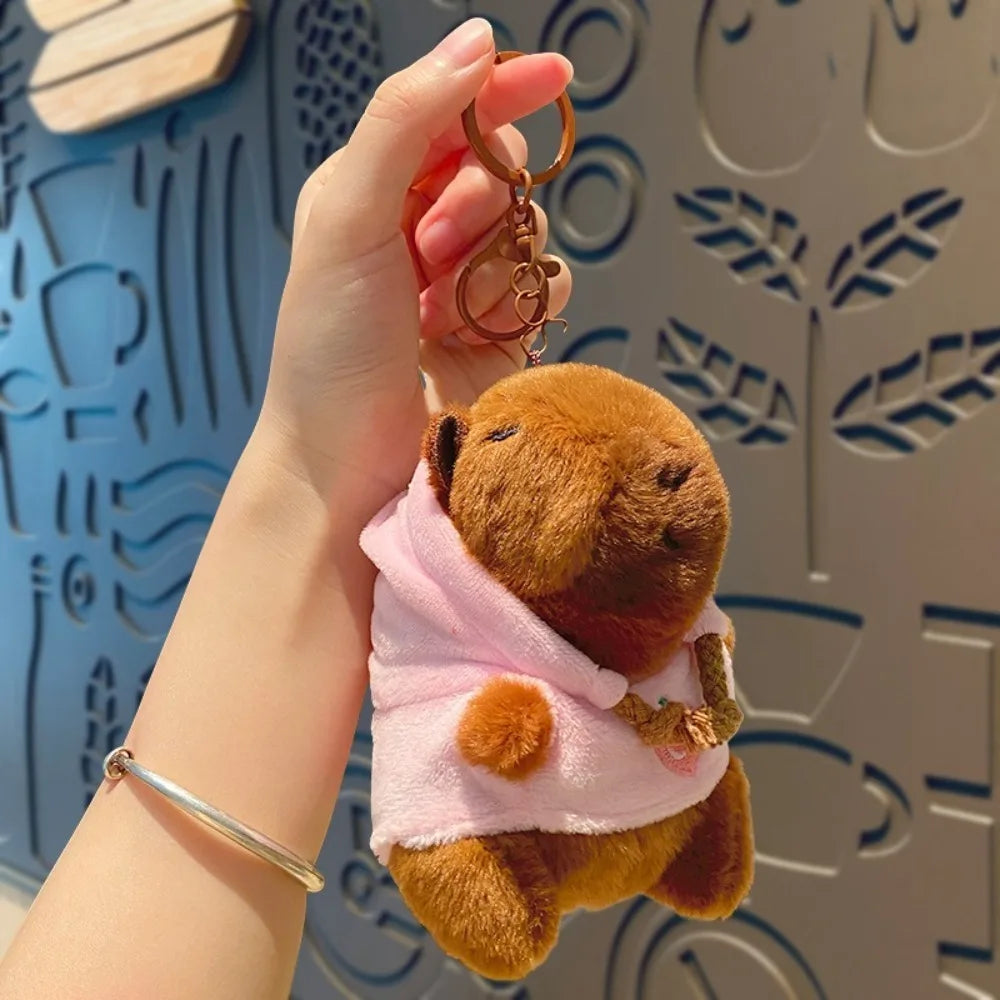 A hand holds a brown capybara plush doll wearing a pink hoodie and gold wings against a light blue patterned background.