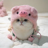 Flauschige rosa Schwein-förmige Mütze, getragen von einer kleinen Katze auf weißem Stoff.
