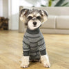 A small dog wearing a gray and white striped jumpsuit standing indoors on a wooden floor.