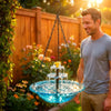 A man is holding a solar-powered hanging bird bath fountain with a glass bowl outdoors.