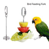Tenaza de acero inoxidable para alimentar pájaros con gancho, junto a frutas y verduras, incluyendo un loro amarillo.
