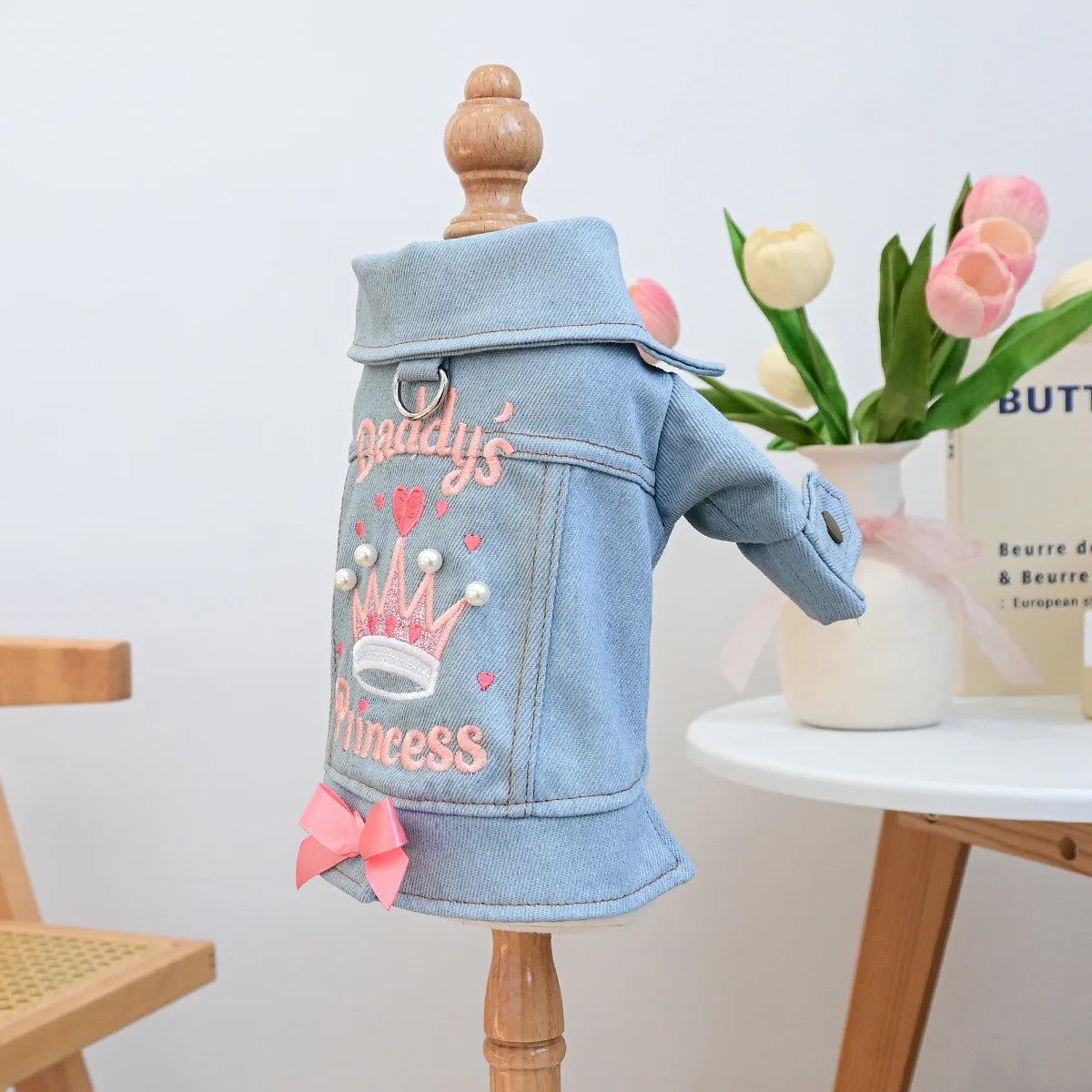 Light blue denim jacket with a crown embroidery and pink bow displayed on a wooden hanger.