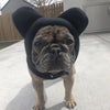 A fawn-colored dog wearing a black fleece winter hat with large ears, standing outdoors.