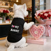 A white dog wearing a black vest with 'I love my Mommy' written on it, sitting in a bright indoor setting.