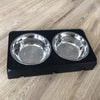A black adjustable raised dog feeder with two stainless steel bowls resting on a light wood surface.